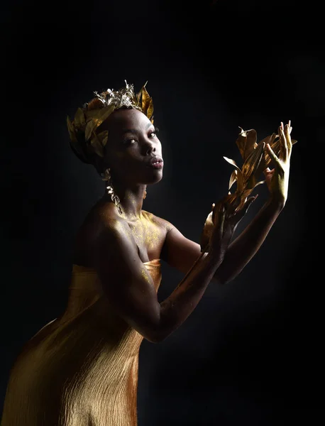 fantasy portrait of beautiful african woman model with afro, goddess silk robes and ornate floral wreath crown. gestural Posing holding golden flowers. isolated on dark  studio background
