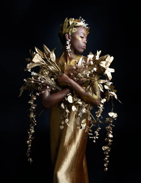 fantasy portrait of beautiful african woman model with afro, goddess silk robes and ornate floral wreath crown. gestural Posing holding golden flowers. isolated on dark  studio background