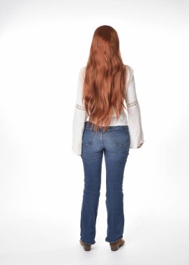Uzun kızıl saçlı güzel bir kadın modelin tam boy portresi, beyaz bluz ve kot pantolon giyiyor, beyaz stüdyo arka planında izole edilmiş. Geriye doğru duruş, kameradan uzaklaşma..