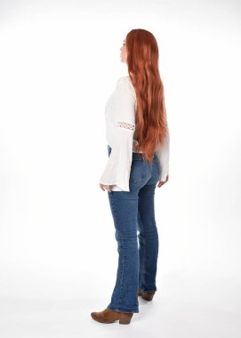 Uzun kızıl saçlı güzel bir kadın modelin tam boy portresi, beyaz bluz ve kot pantolon giyiyor, beyaz stüdyo arka planında izole edilmiş. Geriye doğru duruş, kameradan uzaklaşma..