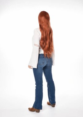 Uzun kızıl saçlı güzel bir kadın modelin tam boy portresi, beyaz bluz ve kot pantolon giyiyor, beyaz stüdyo arka planında izole edilmiş. Geriye doğru duruş, kameradan uzaklaşma..