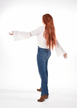 Uzun kızıl saçlı güzel bir kadın modelin tam boy portresi, beyaz bluz ve kot pantolon giyiyor, beyaz stüdyo arka planında izole edilmiş. Geriye doğru duruş, kameradan uzaklaşma..