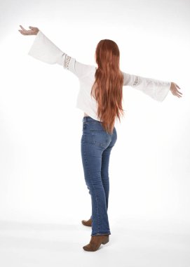 Uzun kızıl saçlı güzel bir kadın modelin tam boy portresi, beyaz bluz ve kot pantolon giyiyor, beyaz stüdyo arka planında izole edilmiş. Geriye doğru duruş, kameradan uzaklaşma..