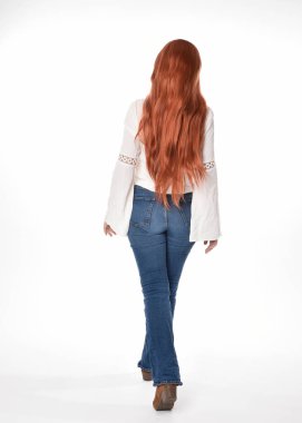 Uzun kızıl saçlı güzel bir kadın modelin tam boy portresi, beyaz bluz ve kot pantolon giyiyor, beyaz stüdyo arka planında izole edilmiş. Geriye doğru duruş, kameradan uzaklaşma..