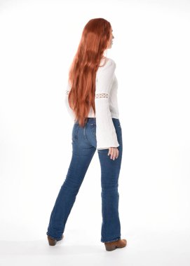 Uzun kızıl saçlı güzel bir kadın modelin tam boy portresi, beyaz bluz ve kot pantolon giyiyor, beyaz stüdyo arka planında izole edilmiş. Geriye doğru duruş, kameradan uzaklaşma..