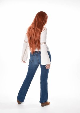 Uzun kızıl saçlı güzel bir kadın modelin tam boy portresi, beyaz bluz ve kot pantolon giyiyor, beyaz stüdyo arka planında izole edilmiş. Geriye doğru duruş, kameradan uzaklaşma..
