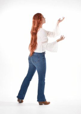 Uzun kızıl saçlı güzel bir kadın modelin tam boy portresi, beyaz bluz ve kot pantolon giyiyor, beyaz stüdyo arka planında izole edilmiş. Geriye doğru duruş, kameradan uzaklaşma..