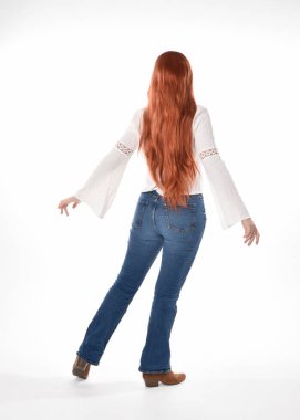 Uzun kızıl saçlı güzel bir kadın modelin tam boy portresi, beyaz bluz ve kot pantolon giyiyor, beyaz stüdyo arka planında izole edilmiş. Geriye doğru duruş, kameradan uzaklaşma..