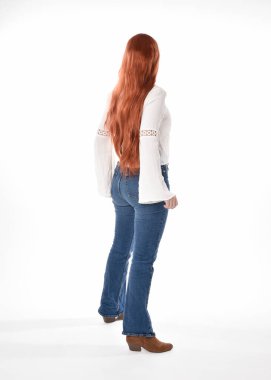 Uzun kızıl saçlı güzel bir kadın modelin tam boy portresi, beyaz bluz ve kot pantolon giyiyor, beyaz stüdyo arka planında izole edilmiş. Geriye doğru duruş, kameradan uzaklaşma..