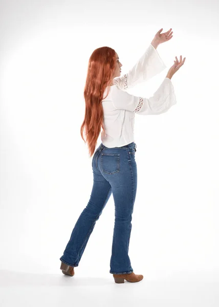 長い赤い髪の美しい女性モデルの完全な長さの肖像画 白いスタジオの背景に隔離されたカジュアル服の白いブラウスの上とデニムジーンズを身に着けている 立ちポーズ カメラから離れて歩く — ストック写真