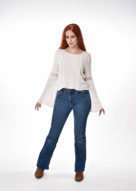 Uzun kızıl saçlı güzel bir kadın modelin tam boy portresi, beyaz bluz ve kot pantolon giyiyor, beyaz stüdyo arka planında izole edilmiş. Duruş, kameraya doğru hareketli el pozları..