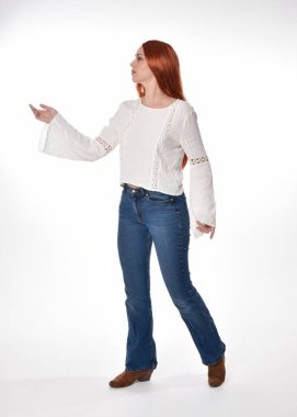 Uzun kızıl saçlı güzel bir kadın modelin tam boy portresi, beyaz bluz ve kot pantolon giyiyor, beyaz stüdyo arka planında izole edilmiş. Duruş, kameraya doğru hareketli el pozları..