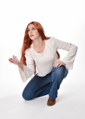 Uzun kızıl saçlı güzel bir kadın modelin tam boy portresi, beyaz bluz ve kot pantolon giyiyor, beyaz stüdyo arka planında izole edilmiş. Rahat bir poz, yerde el hareketleriyle oturuyor..