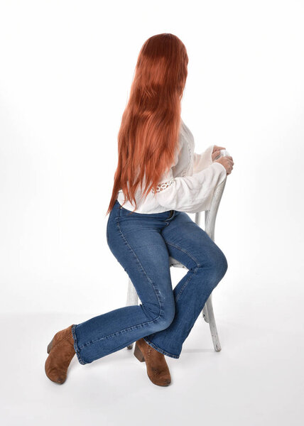 full length portrait of beautiful woman model with long red hair, wearing casual outfit white blouse  top and denim jeans, isolated on white studio background. Relaxed seated pose, sitting on chair with gestural hand poses.