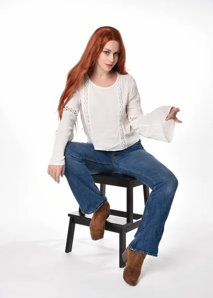 stock image full length portrait of beautiful woman model with long red hair, wearing casual outfit white blouse  top and denim jeans, isolated on white studio background. Relaxed seated pose, sitting on floor with gestural hand poses.