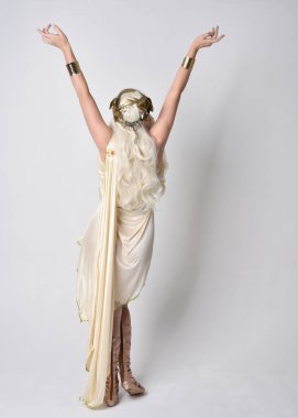 Fantezi tanrıça Toga kostümü giyen güzel sarışın kadının uzun bir portresi. Büyülü bir taç takmış. Duruş alıyor, kameradan geriye bakıyor. Beyaz stüdyo arka planında izole.