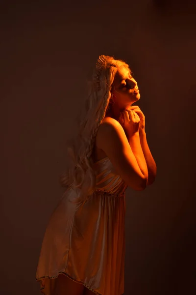 身着奇幻女神托加服装 头戴水晶王冠的金发美女的肖像 背对着一个闪烁着橙色轮廓灯光的发光的独立工作室背景 — 图库照片