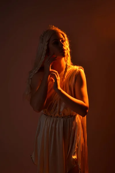身着奇幻女神托加服装 头戴水晶王冠的金发美女的肖像 背对着一个闪烁着橙色轮廓灯光的发光的独立工作室背景 — 图库照片