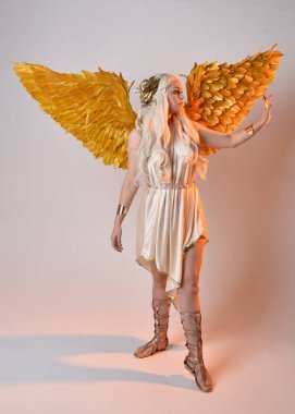Tüylü melek kanatları olan fantezi tanrıça toga kostümü giyen güzel sarışın kadının tam boy portresi. Atlama pozu, beyaz stüdyo arka planında izole edilmiş..