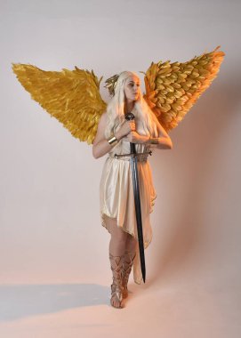Uzun boylu, sarışın güzel bir kadının fantezi tanrıça toga kostümü giymiş tüylü melek kanatları olan, kılıç tutan bir kadın portresi. Atlama pozu, beyaz stüdyo arka planında izole edilmiş..