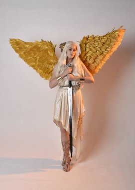 Uzun boylu, sarışın güzel bir kadının fantezi tanrıça toga kostümü giymiş tüylü melek kanatları olan, kılıç tutan bir kadın portresi. Atlama pozu, beyaz stüdyo arka planında izole edilmiş..