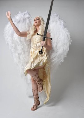 Uzun boylu, sarışın güzel bir kadının fantezi tanrıça toga kostümü giymiş tüylü melek kanatları olan, kılıç tutan bir kadın portresi. Atlama pozu, beyaz stüdyo arka planında izole edilmiş..