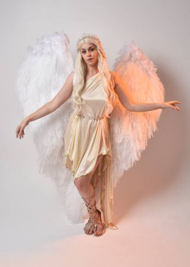 Tüylü melek kanatları olan fantezi tanrıça toga kostümü giyen güzel sarışın kadının tam boy portresi. Atlama pozu, beyaz stüdyo arka planında izole edilmiş..