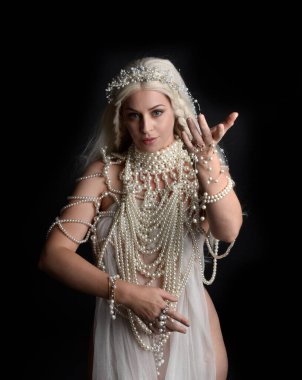 Güzel sarışın mankenin, abartılı bir inci mücevher elbisesi giymiş portresi. Jeolojik kollar uzanıyor, siyah bir stüdyoda izole edilmiş..