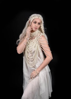 Güzel sarışın mankenin, abartılı bir inci mücevher elbisesi giymiş portresi. Jeolojik kollar uzanıyor, siyah bir stüdyoda izole edilmiş..