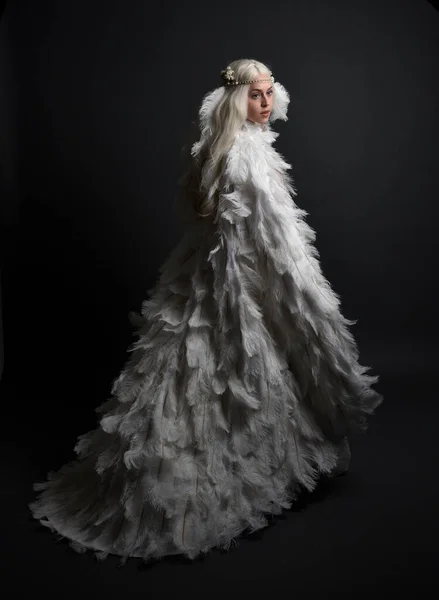 stock image fantasy portrait of beautiful female model with long blond hair wearing otherworldly  white feathered cloak costume and headdress, isolated on dark studio background.
