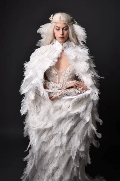 stock image fantasy portrait of beautiful female model with long blond hair wearing otherworldly  white feathered cloak costume and headdress, isolated on dark studio background.