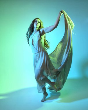 Güzel esmer kadın dansçının uzun bir portresi, ruhani bir elbise giyiyor, akıcı kumaşla dans ediyor. Sinema renkli ışıklandırmayla yeşil stüdyo arka planında izole edilmiş.
