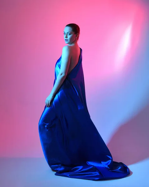 stock image Full length portrait of beautiful female model wearing elegant flowing toga gown of blue silk drapery, isolated on studio background with cinematic colourful lighting.