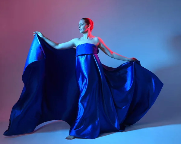 stock image Full length portrait of beautiful female model wearing elegant flowing toga gown of blue silk drapery, isolated on studio background with cinematic colourful lighting.