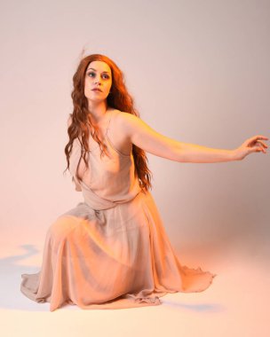 Güzel esmer kadın modelin tam boy portresi altın arka ışıklandırmalı elbise giyiyor. El hareketleriyle diz çökme pozu. Stüdyo arka planında izole.