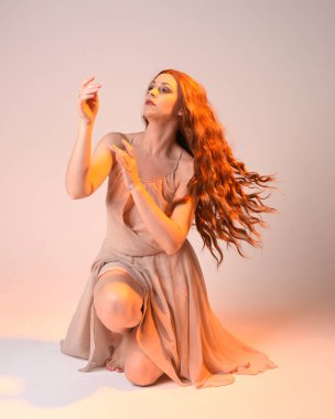 Güzel esmer kadın modelin tam boy portresi altın arka ışıklandırmalı elbise giyiyor. El hareketleriyle diz çökme pozu. Stüdyo arka planında izole.