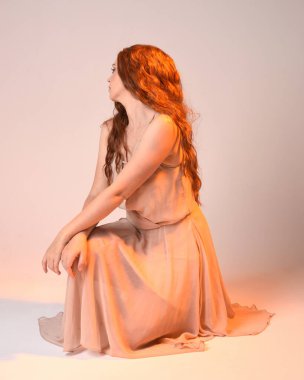 Güzel esmer kadın modelin tam boy portresi altın arka ışıklandırmalı elbise giyiyor. El hareketleriyle diz çökme pozu. Stüdyo arka planında izole.