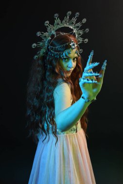 Süslü gümüş taç başlık takan güzel esmer kadının portresi, kol hareketleriyle poz vermek, sihirli büyü yapmak. Karanlık stüdyo arka planında izole edilmiş, sinematik mavi ay ışığı.