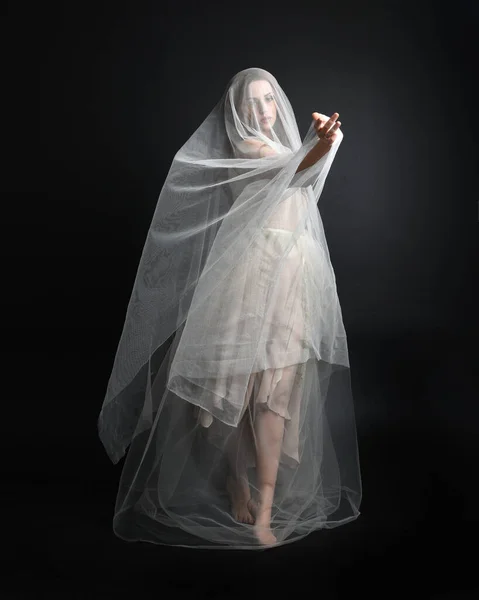 ダークスタジオのバックグラウンドで隔離された幽霊に覆われた生地で白いガウンのドレスを着た美しい女性の完全な長さの肖像画 — ストック写真