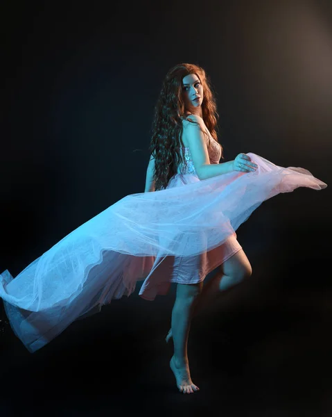 stock image Full length  portrait of beautiful brunette woman dancer, wearing ethereal gown, dancing with flowing fabric.  isolated on dark studio background with cinematic colourful lighting.