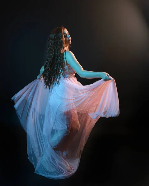 stock image Full length  portrait of beautiful brunette woman dancer, wearing ethereal gown, dancing with flowing fabric.  isolated on dark studio background with cinematic colourful lighting.