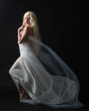 Sarışın güzel bir kadının beyaz elbiseler giydiği uzun bir portre. Örtülü beyaz bir elbise. Dar stüdyo arka planında izole edilmiş..