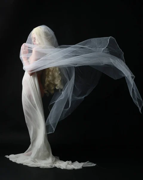 美しいブロンドの女性の完全な長さの肖像画は 暗いスタジオの背景に隔離された幽霊に覆われた生地で白いガウンのドレスを着ています — ストック写真