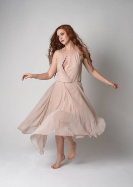 Uzun, esmer saçlı, kremsi pembe bir elbise giyen güzel bir kadın modelin tam boy portresi. Beyaz stüdyo arka planında el hareketleriyle zarif bir dans pozisyonu..