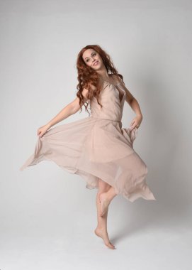 Uzun, esmer saçlı, kremsi pembe bir elbise giyen güzel bir kadın modelin tam boy portresi. Beyaz stüdyo arka planında el hareketleriyle zarif bir dans pozisyonu..