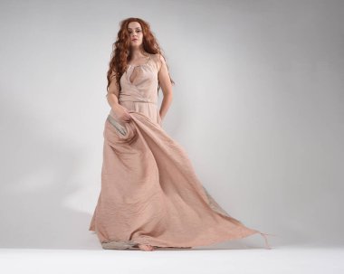 Kremsi pembe bir elbise giyen güzel esmer kadın modelin tam boy portresi. Düşük perspektiften çekilen zarif dans pozu, beyaz stüdyo arka planında el hareketleriyle izole edilmiş..