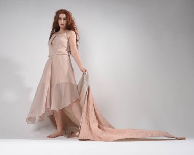 Kremsi pembe bir elbise giyen güzel esmer kadın modelin tam boy portresi. Düşük perspektiften çekilen zarif dans pozu, beyaz stüdyo arka planında el hareketleriyle izole edilmiş..