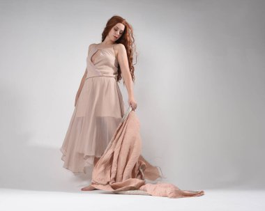 Kremsi pembe bir elbise giyen güzel esmer kadın modelin tam boy portresi. Düşük perspektiften çekilen zarif dans pozu, beyaz stüdyo arka planında el hareketleriyle izole edilmiş..