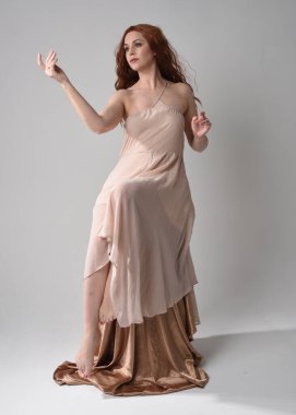 Uzun, esmer saçlı, kremsi pembe bir elbise giyen güzel bir kadın modelin tam boy portresi. Beyaz stüdyo arka planında izole edilmiş zarif oturma pozu.