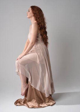 Uzun, esmer saçlı, kremsi pembe bir elbise giyen güzel bir kadın modelin tam boy portresi. Beyaz stüdyo arka planında izole edilmiş zarif oturma pozu.